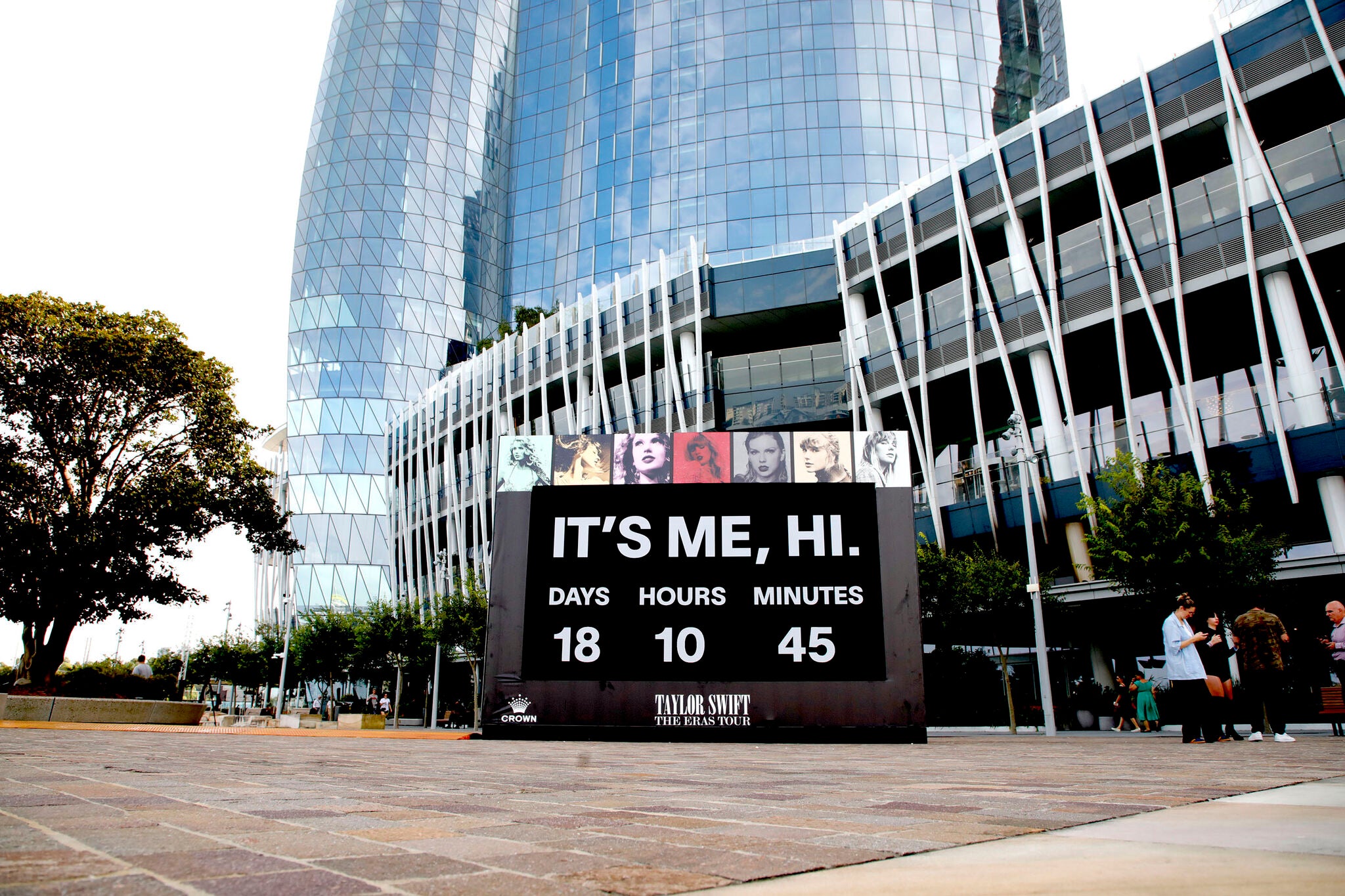 Digital clock displaying 'It's Me, Hi.' in front of a modern building with people walking by for Taylor Swift Eras Tour