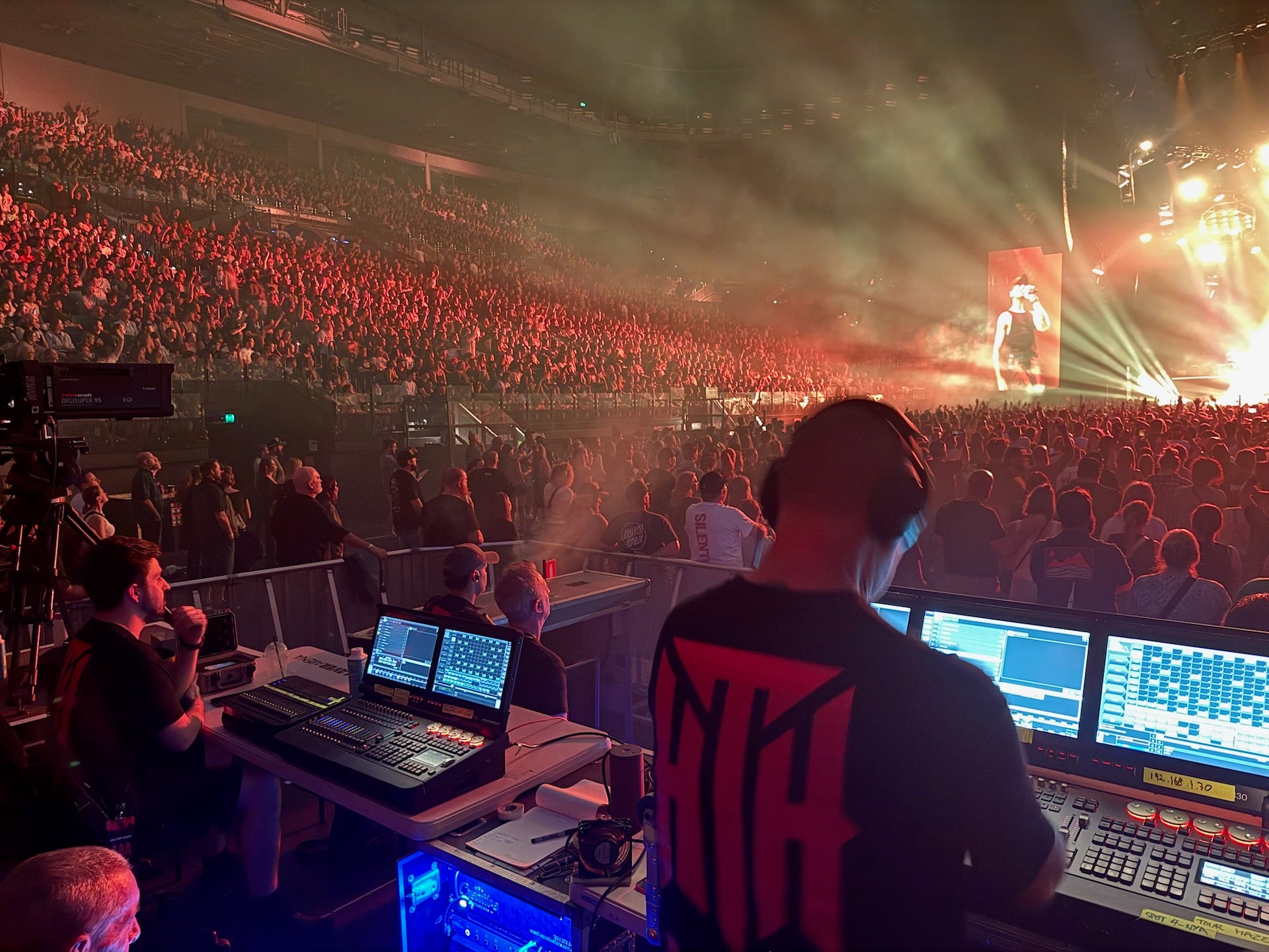 Control room with multiple monitors and people monitoring a large event with a crowd and stage lights.