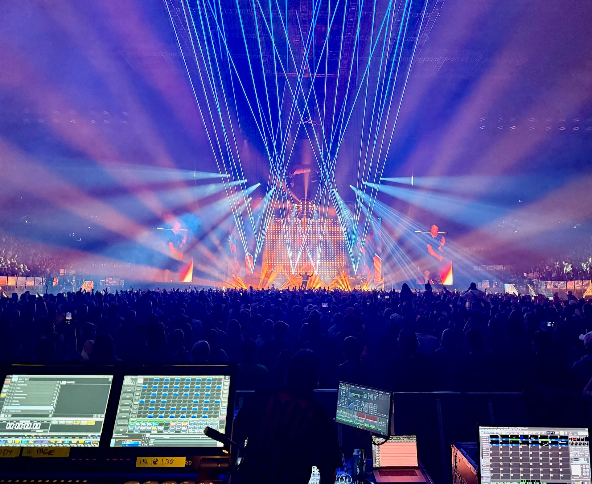 Dj booth with screens and equipment during a concert with colorful laser lights and stage.