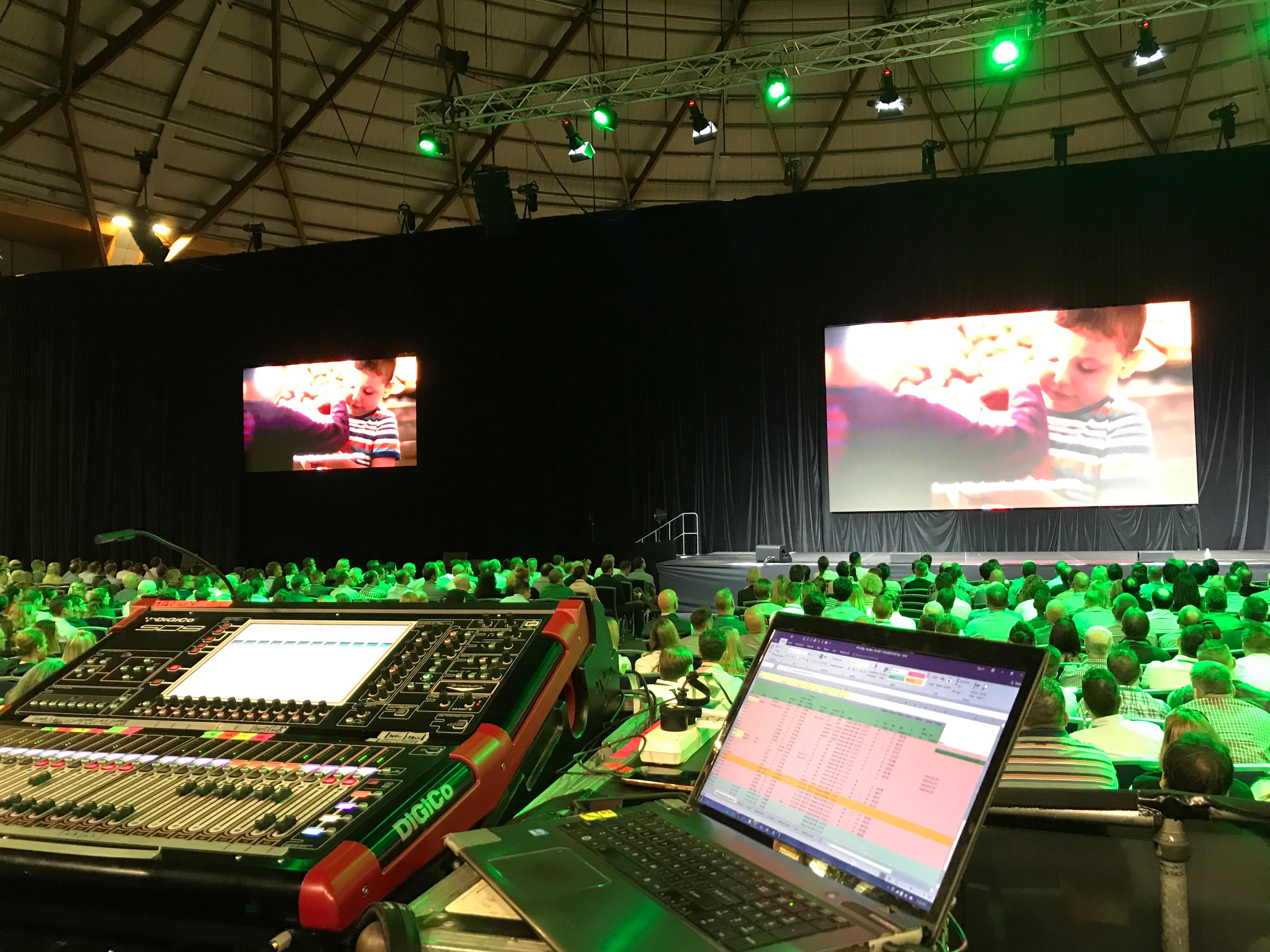 Control room with audio mixing console and laptop displaying software, large screens showing a person working on a computer at a conference