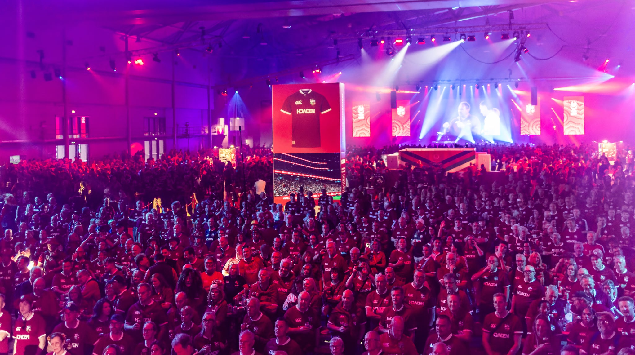 Large indoor event with a stage, large screen, and enthusiastic crowd.