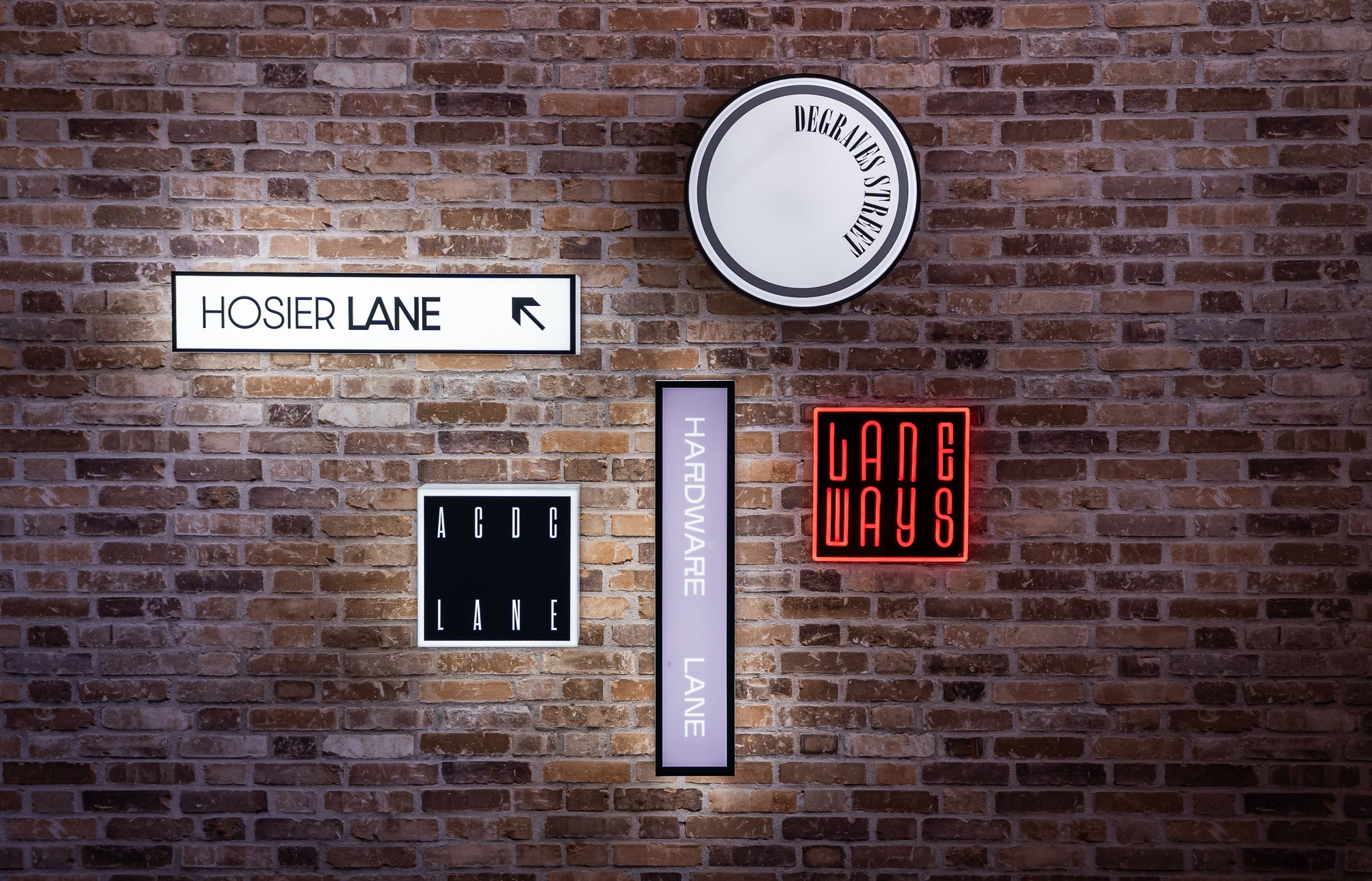 Neon signs on a faux brick wall with text like 'Hosier Lane' and 'Lane Ways'. Custom illuminated signage at Australian Grand Prix