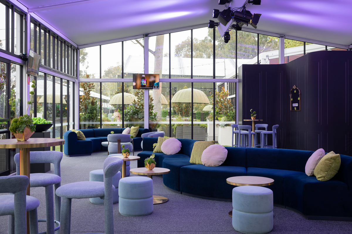 A modern lounge area with curved dark blue sofas, light blue cylindrical ottomans, and gray chairs in a bright room with large windows and purple ambient lighting.