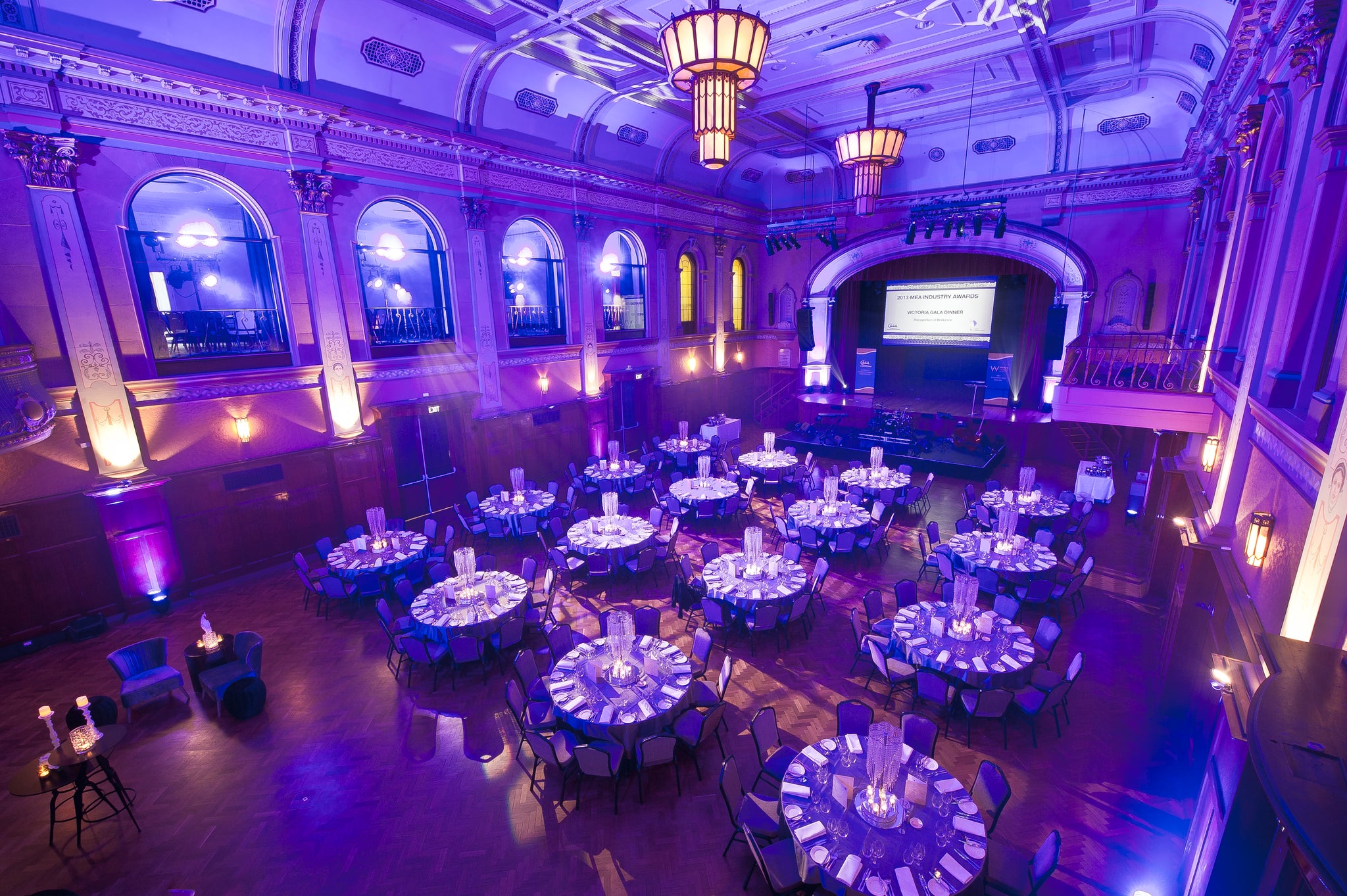 Grand ballroom with round tables set for an event, illuminated with purple and blue lights.