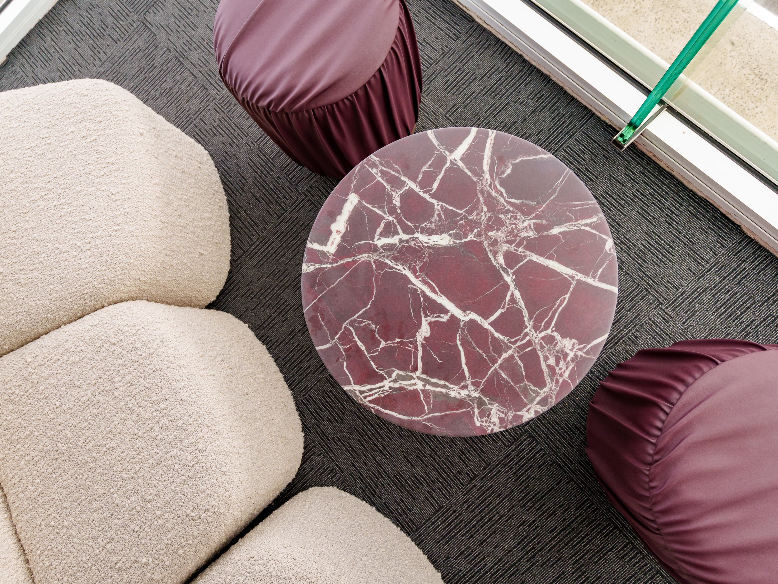 Round marble Halo table with a beige sofa and maroon Rocco cushioned stools on a dark floor.
