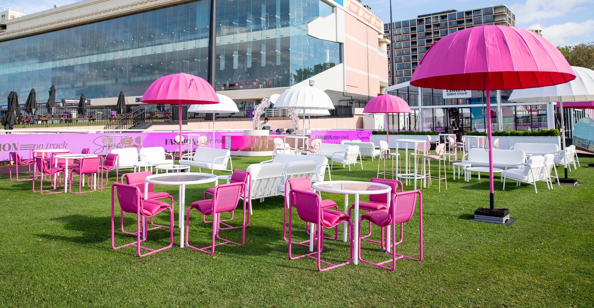 Outdoor setting with Isla pink tables, chairs, and umbrellas on a grassy area with buildings in the background. At Spring Racing Carnival, Melbourne