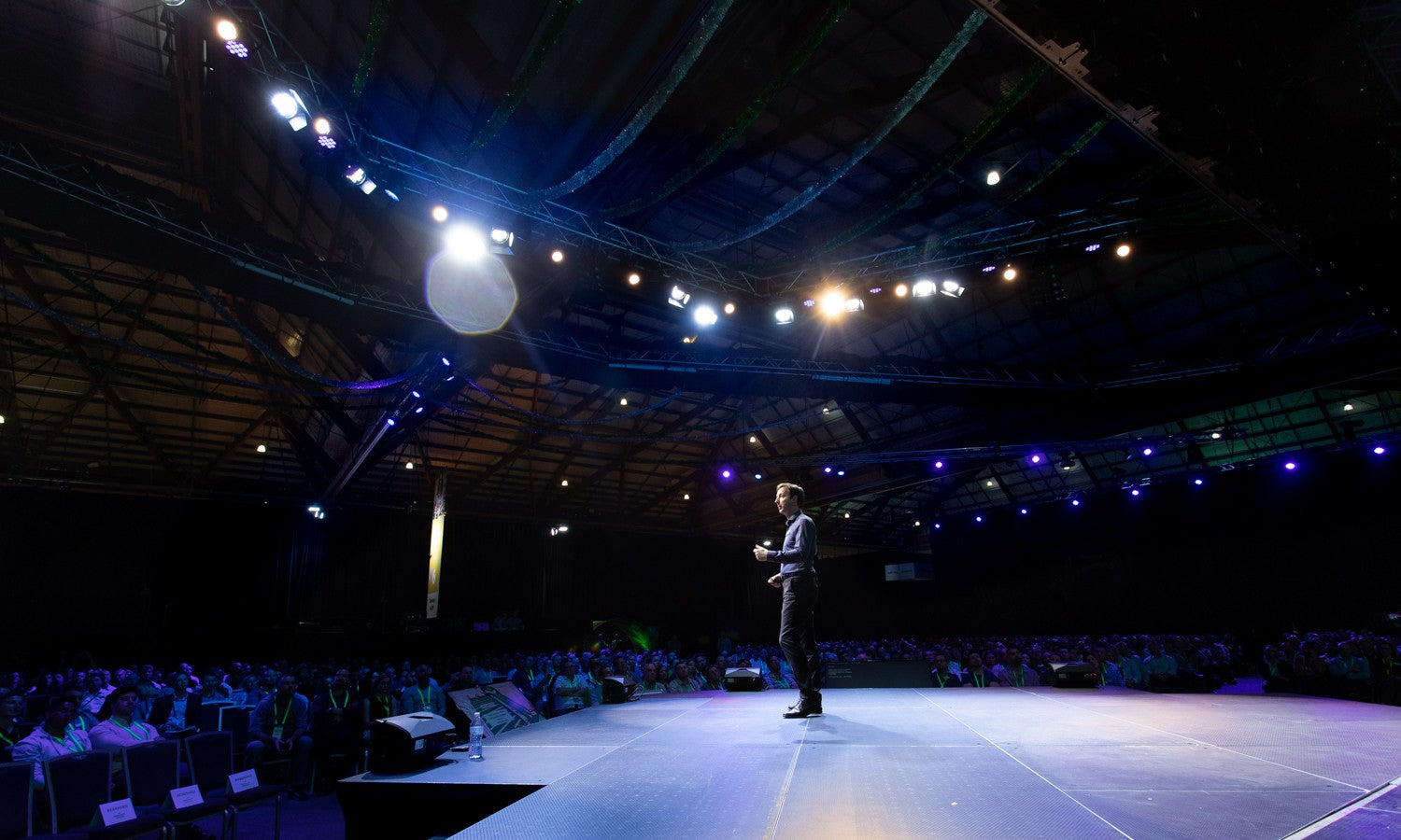 Person standing on a stage in a large auditorium with audience and lighting.