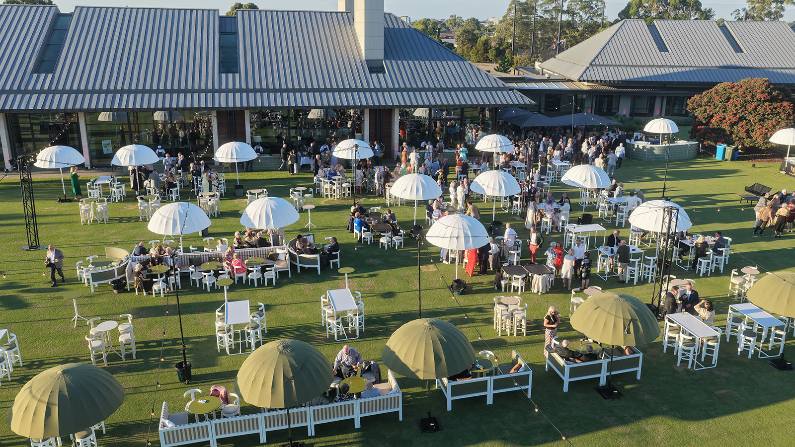 Outdoor event with tables, chairs, and umbrellas on a grassy area with buildings in the background.