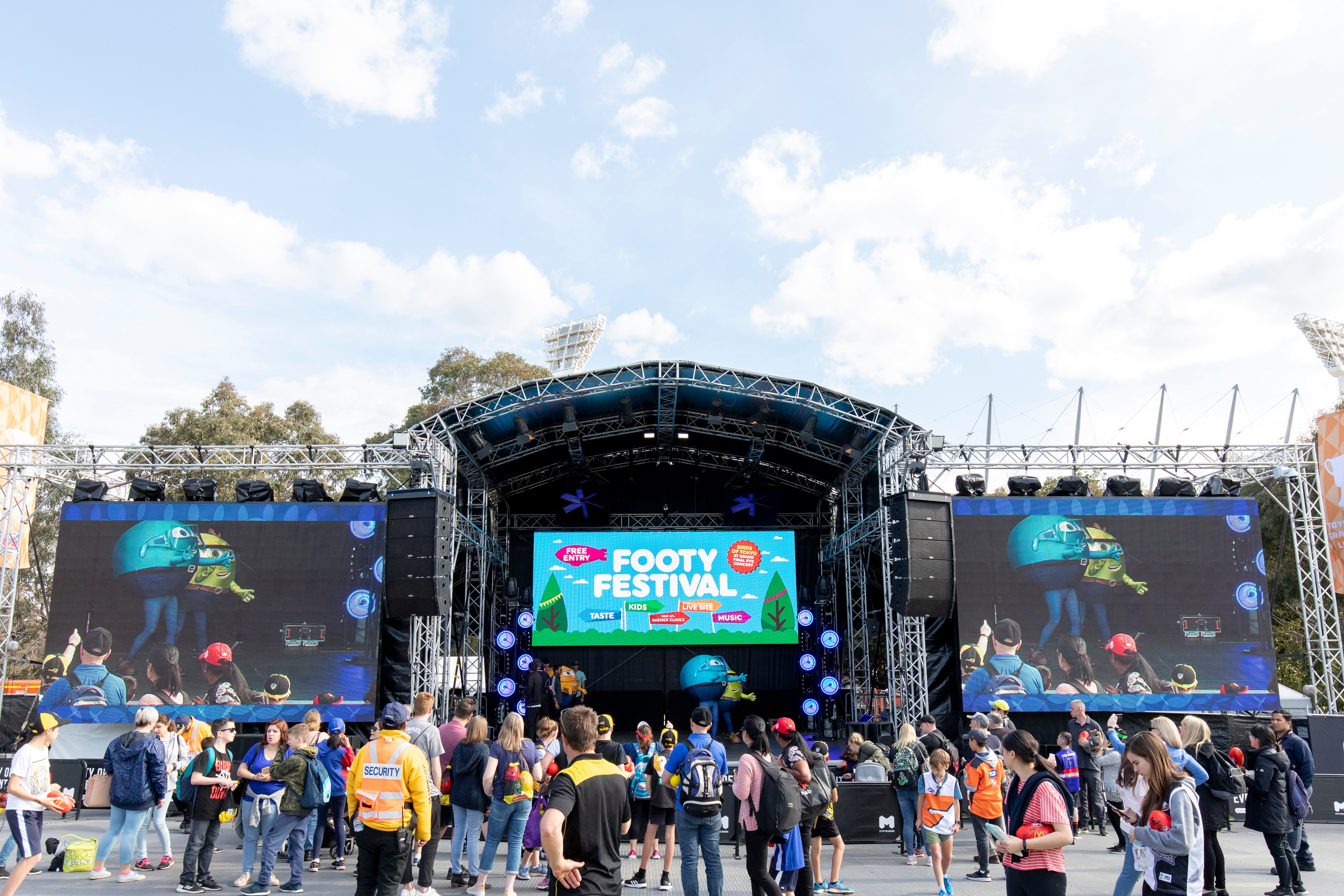 Outdoor event with a rigged stage and large screens displaying 'Footy Festival' on a clear day.