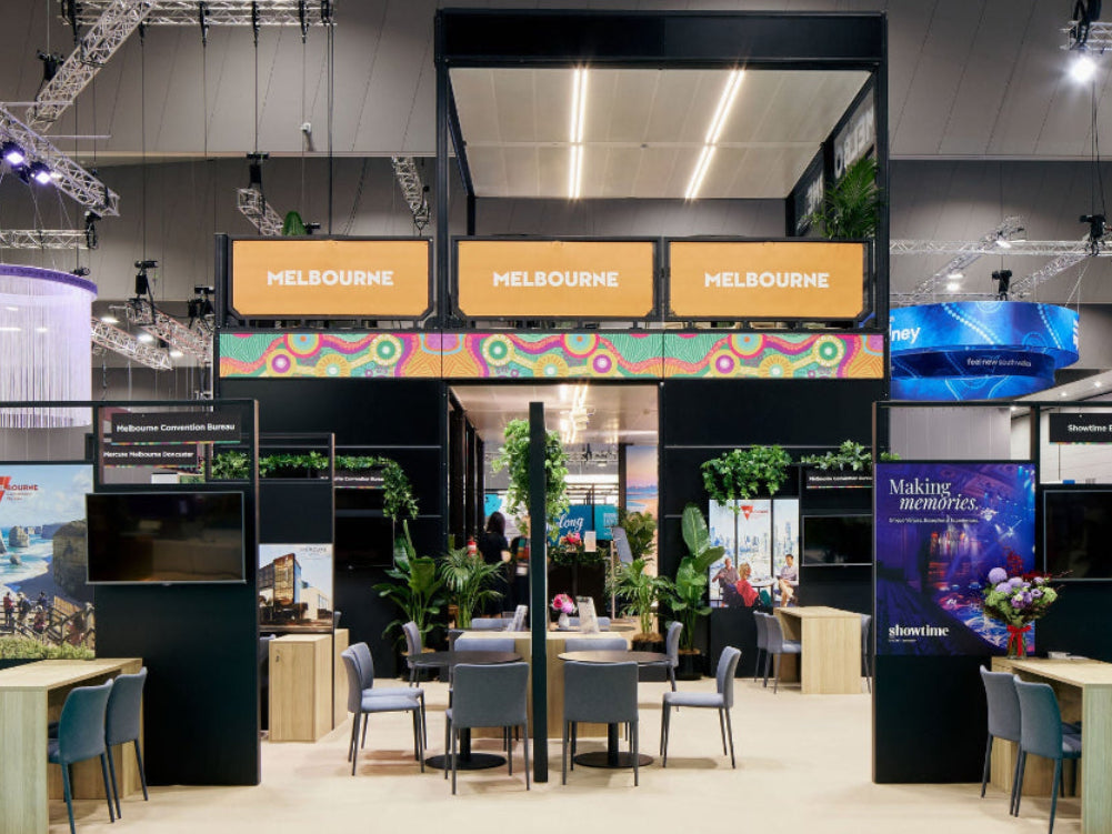 A large exhibition booth at a trade show with the word 'MELBOURNE' repeated across three orange signs above a seating area, tables, chairs, plants, and display screens in a convention hall.