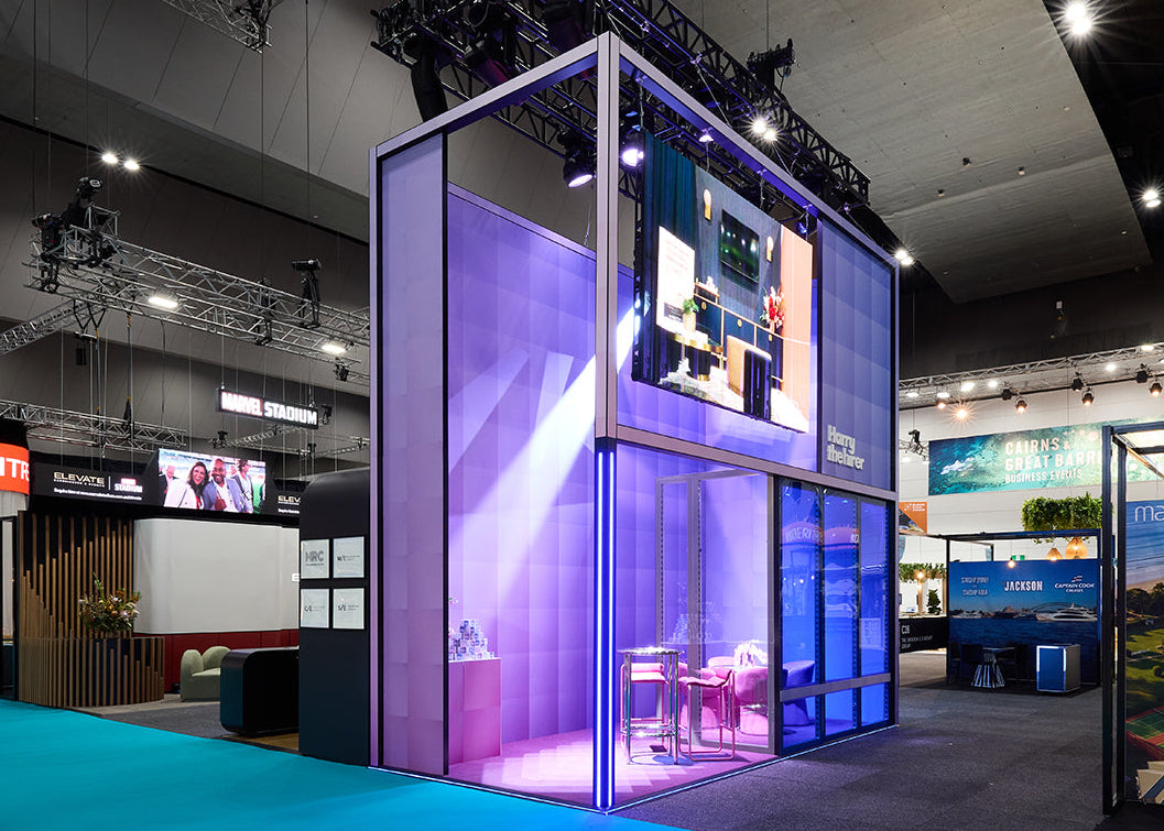 A modern glass booth or exhibition space with purple and blue LED lighting, set up inside a large convention hall. The structure has a white frame, multiple levels, and large display screens on the exterior.