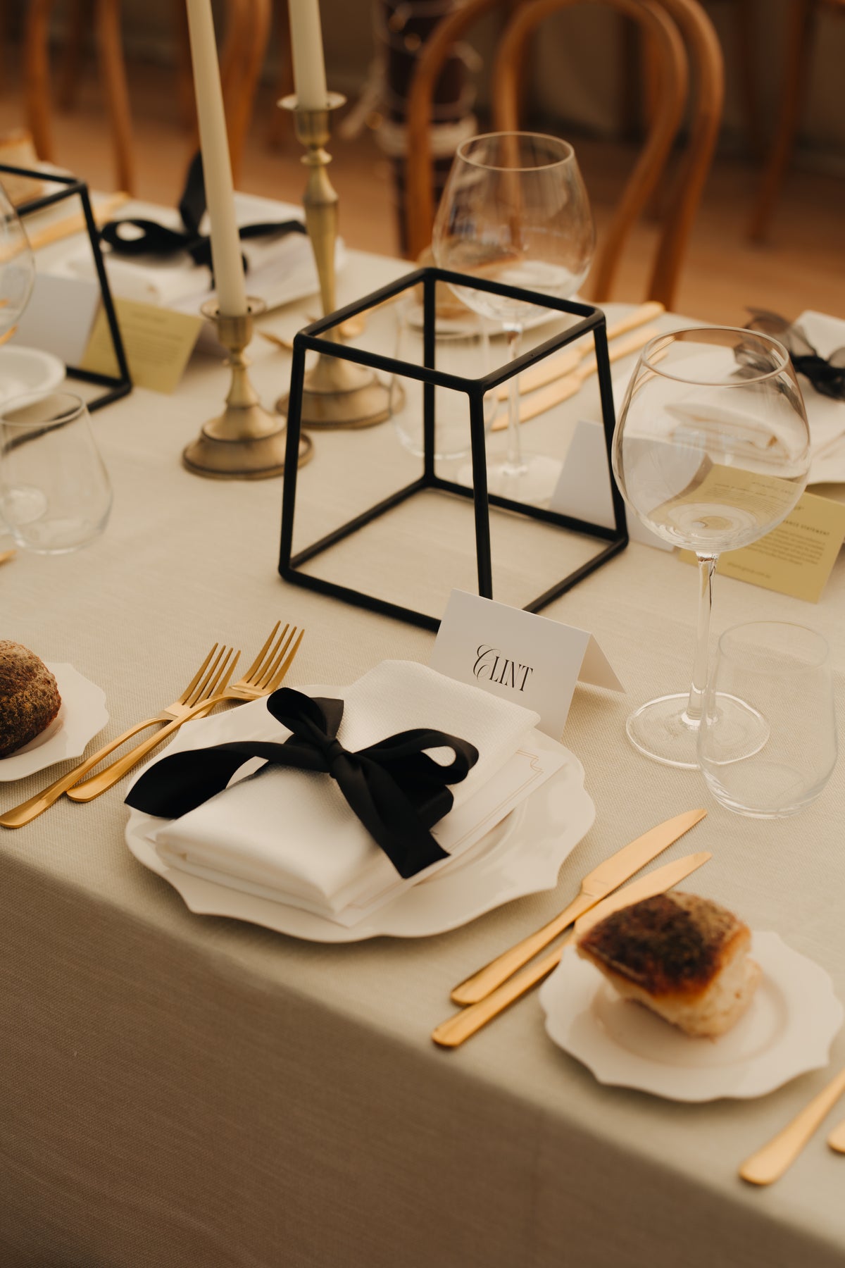 A styled wedding table setting featuring a white napkin tied with a black ribbon, gold flatware, clear wine glasses, and a black geometric wire cube centerpiece on a beige tablecloth.