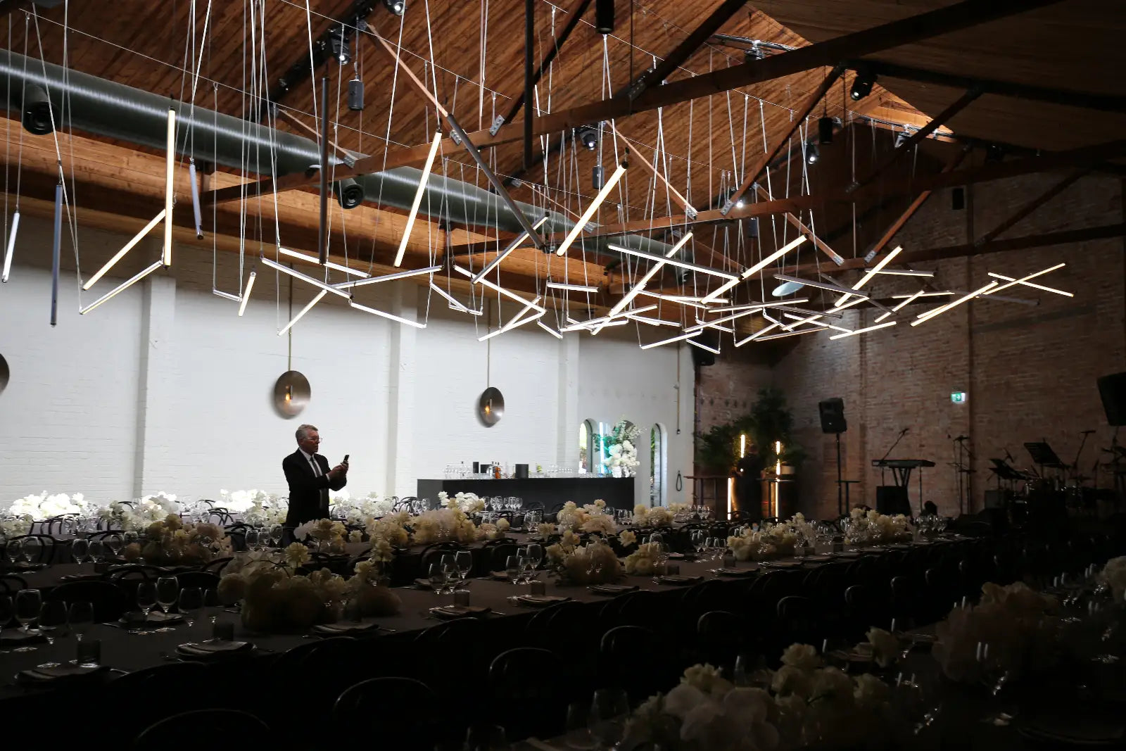 Dining hall with long tables set for a formal event, featuring modern hanging lights and a speaker on stage.