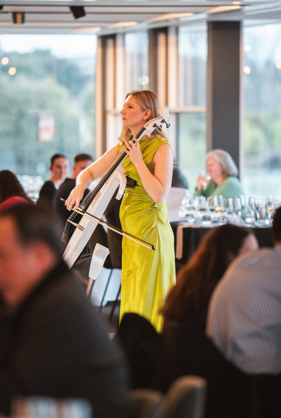 Woman playing a cello in a formal event setting with guests in the background.